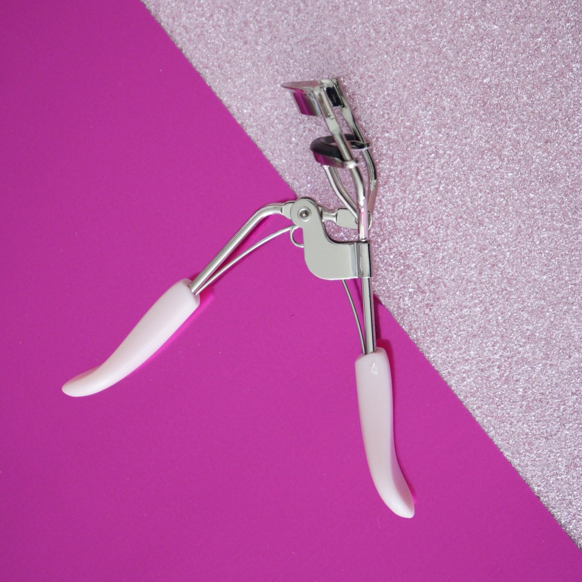 manna kadar pink handle eyelash curler sitting on half pink glitter and solid pink background