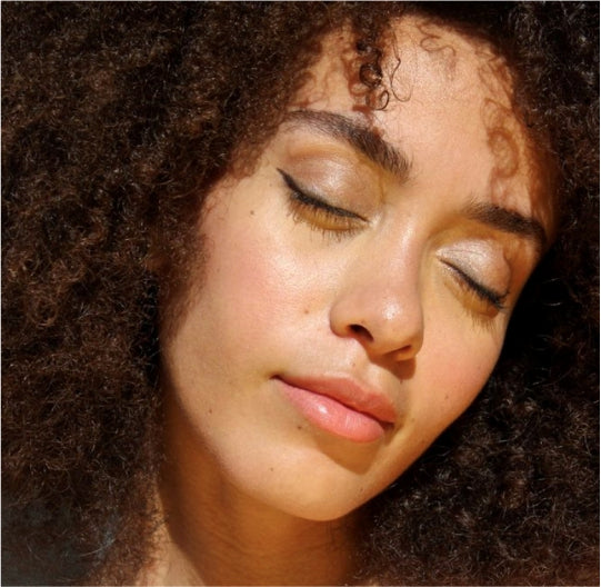 Woman with fluffy curly hair with eyes closed showing off a soft shimmer eyeshadow called Diamond dust by Manna Kadar. Also wearing soft pink and peach gradient blush from Manna Kadar Blossom Blush Compact and a soft natural pink lip using Manna Kadar pH Balance Lip Chemistry lip balm.