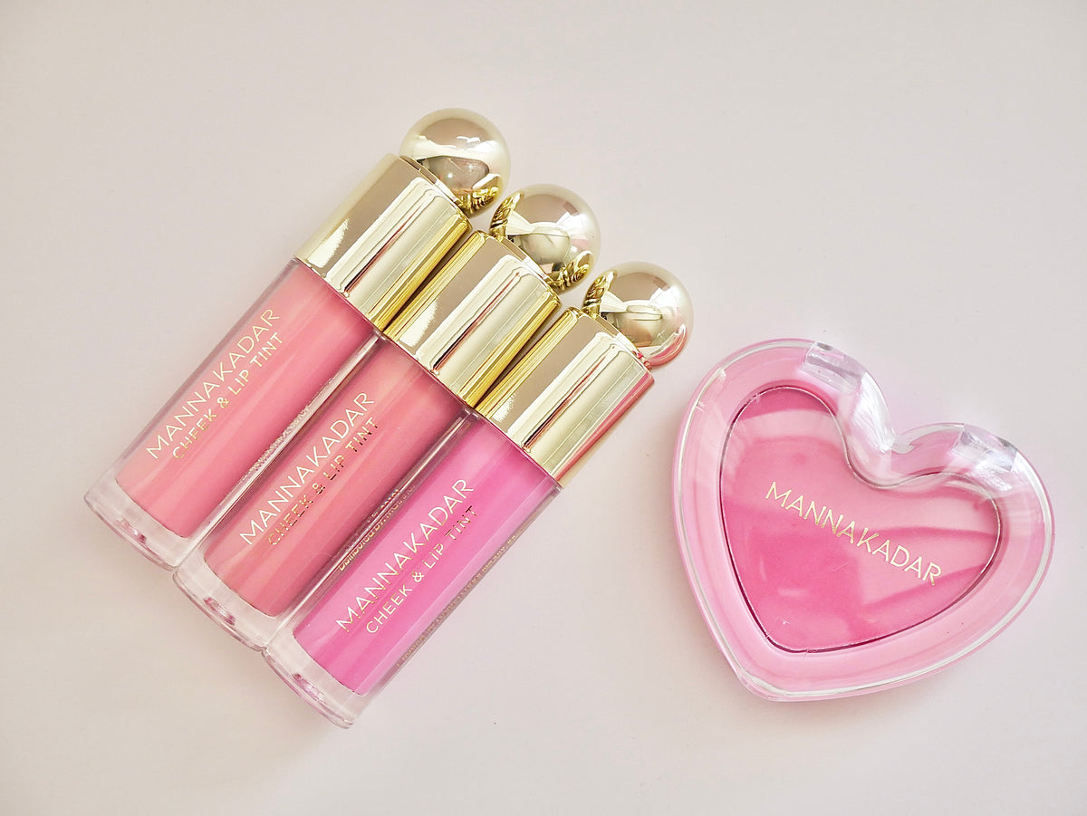 Three pink lip gloss tubes with gold caps arranged next to a pink heart-shaped compact case on a light beige background. The brand name MANA MA KADAR is visible on the products.