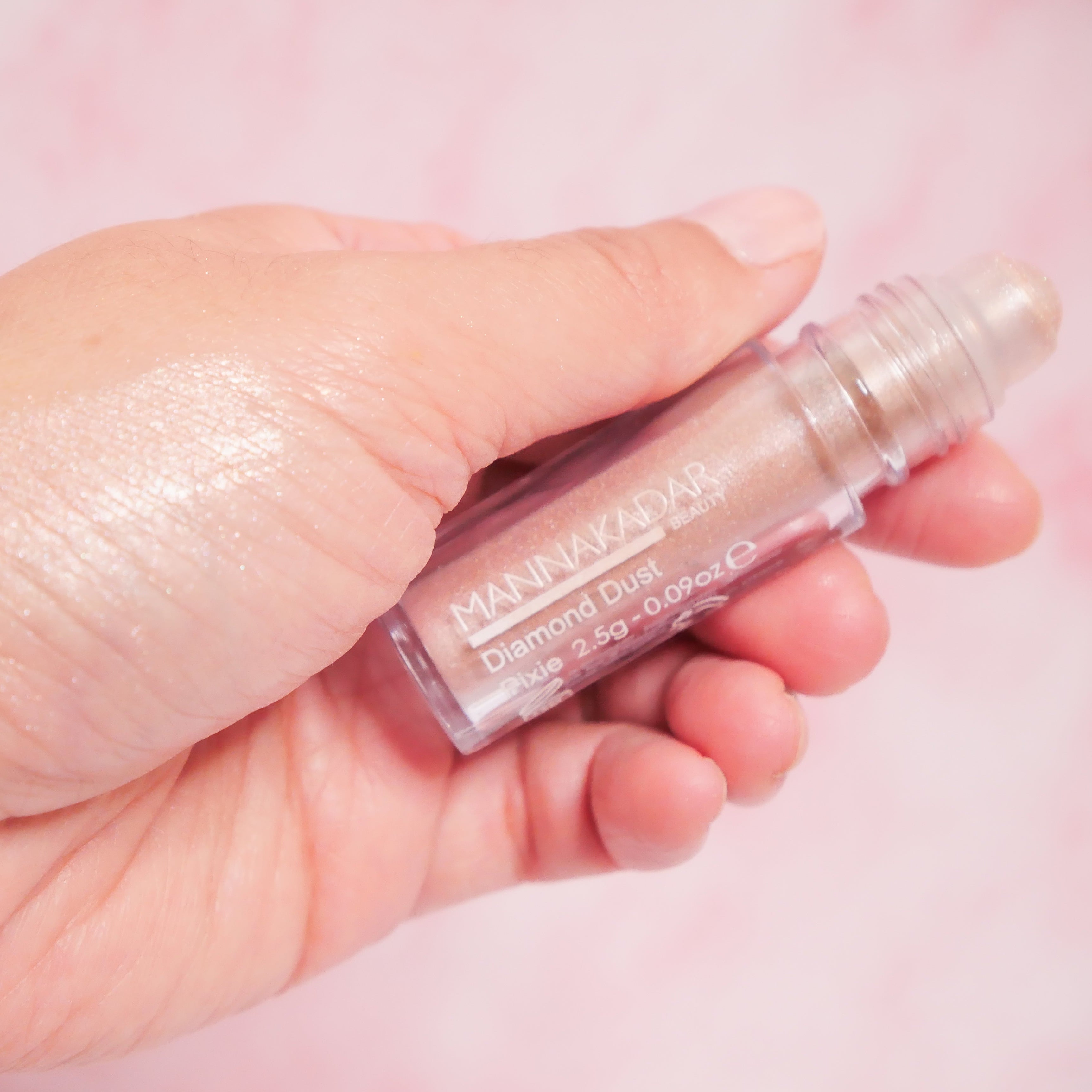 Hand holding a Manic Panic Deluxe Ultra Gloss lip gloss with a pink background