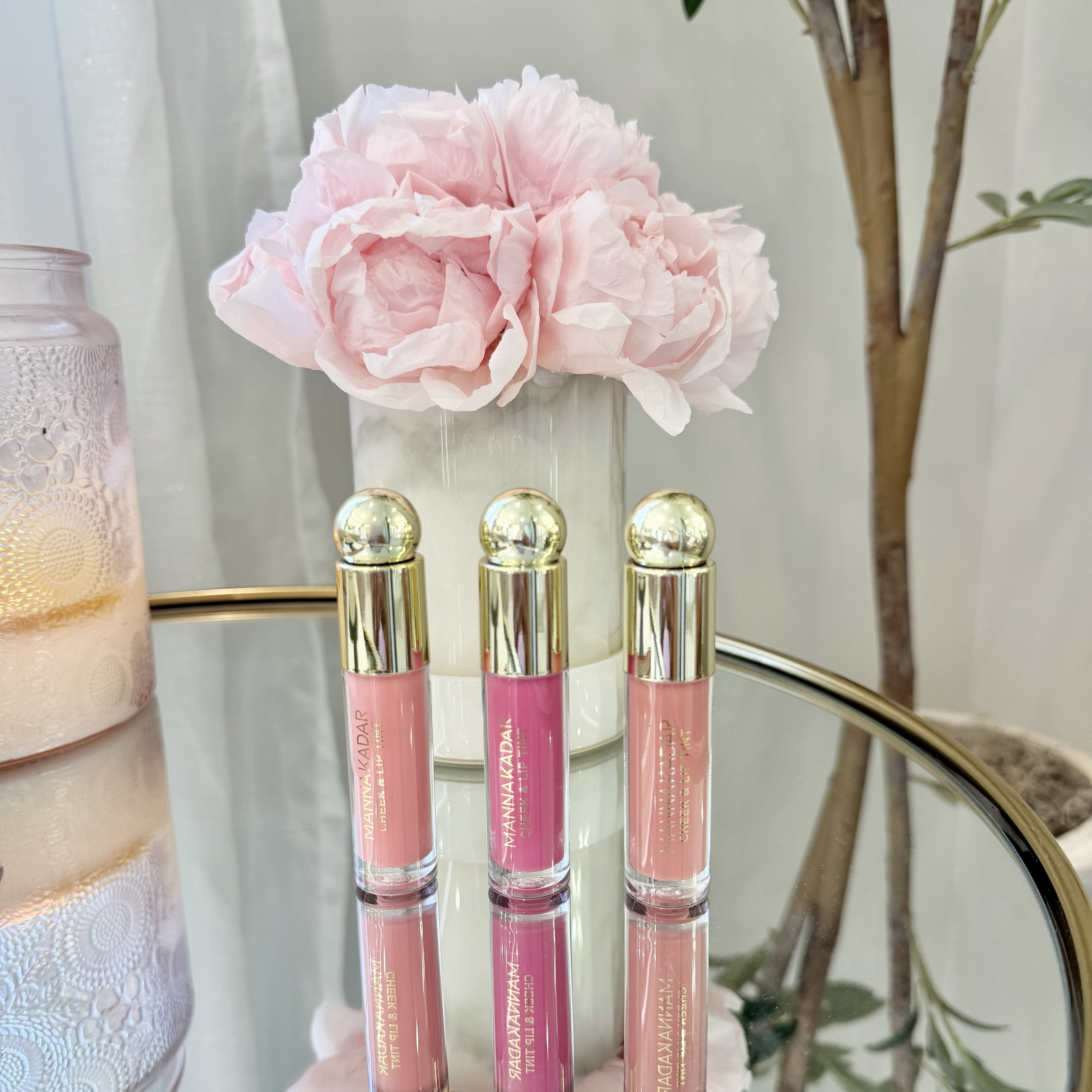Three lip glosses with gold caps on a reflective surface, pink flower in the background