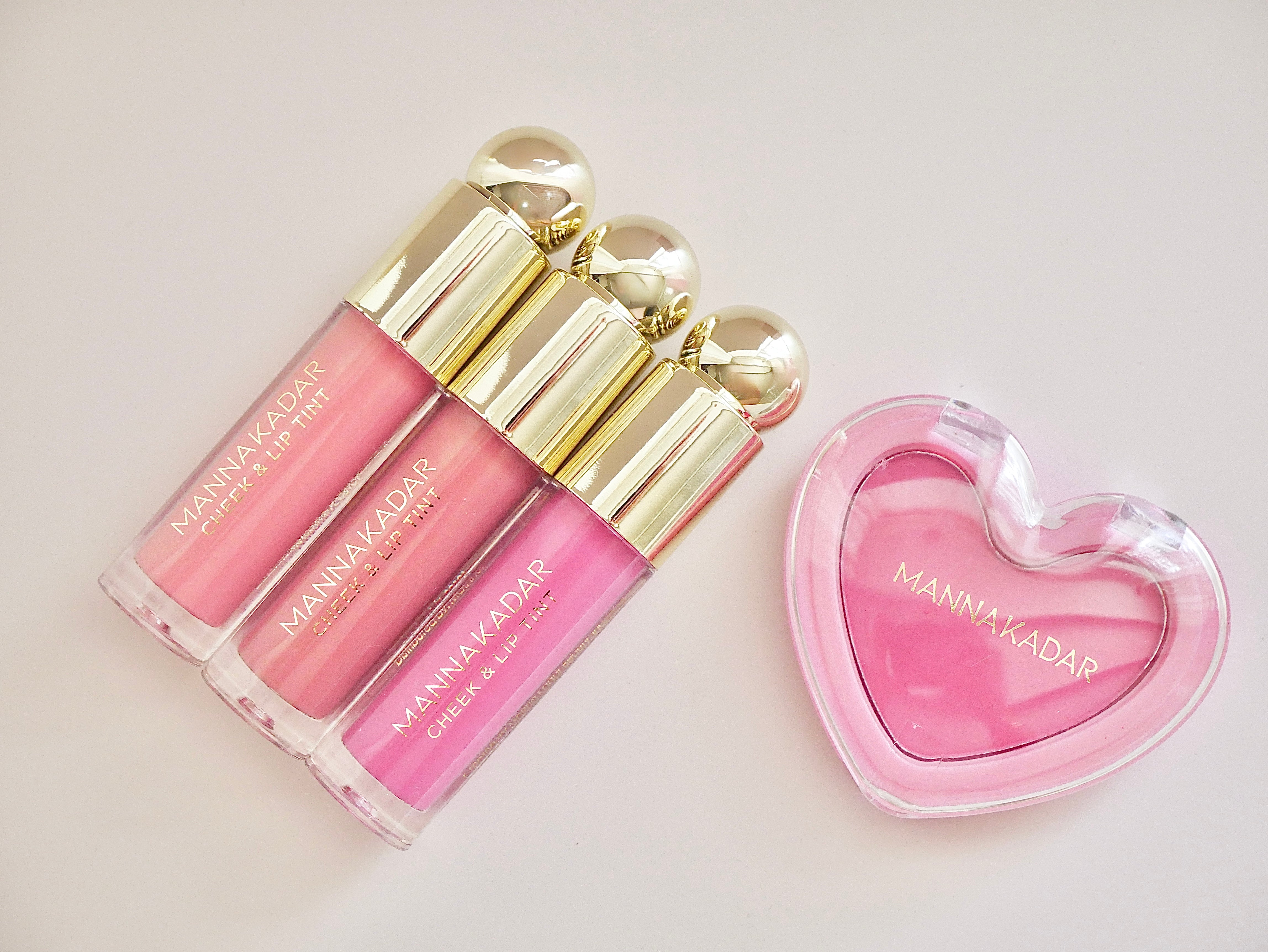 Three pink lip gloss tubes with gold caps arranged next to a pink heart-shaped compact case on a light beige background. The brand name MANA MA KADAR is visible on the products.