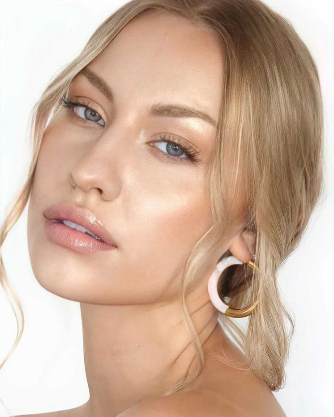 Close-up of a person with long blonde hair, wearing hoop earrings, looking towards the camera against a plain background wearing manna Kadar lip gloss. Makeup is light and clean on fair skin.